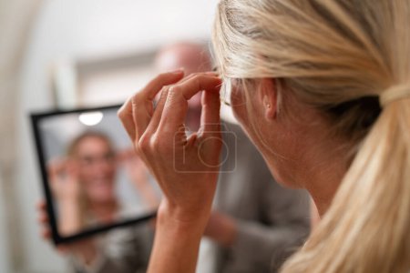 Woman trying on glasses and looking at her reflection in a mirror on a optician store. The focus is on hands holding glasses with blurred background