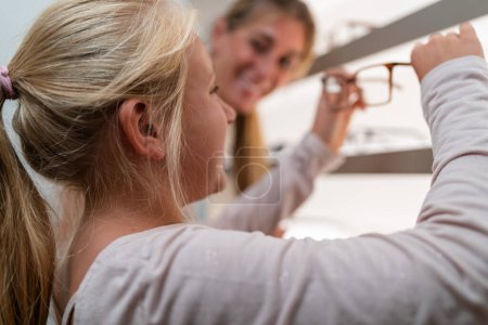 Child reaching for eyeglasses on shelf with adult supervising in optical store to buy new glasses