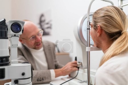 Optometrista ajustando queratografía para un examen ocular en aclínico. Hombre y mujer sentados en una mesa hablando entre sí.