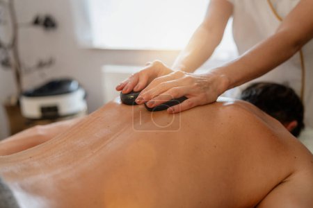 Therapeut gibt einer Klientin, die mit dem Gesicht nach unten liegt, eine Massage mit heißen Steinen