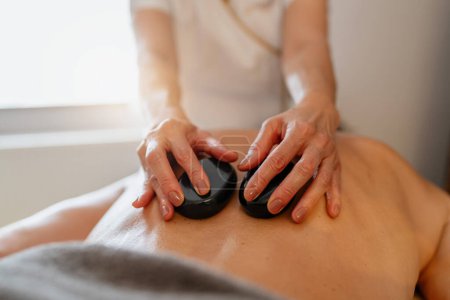 Massagetherapeutin mit heißen Steinen auf dem Rücken des Klienten während einer Wellness-Behandlung. Wellness Hotel Konzept Bild