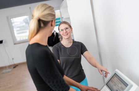 Entraîneuse pointant vers une balance de composition corporelle affiche pour expliquer à son client la fonction d'un test Inbody dans un studio de fitness