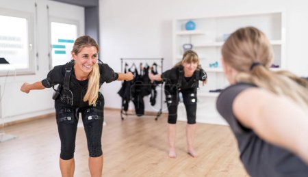 Deux femmes en costume d'entraînement EMS faisant des exercices d'équilibre devant un entraîneur dans une salle de gym