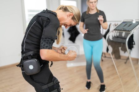 Woman in EMS suit performing exercises with trainer guidance in a gym