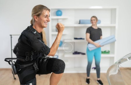 Mujer en traje EMS haciendo sentadillas con entrenador sosteniendo esterilla de yoga para el próximo ejercicio de entrenamiento en un estudio EMS
