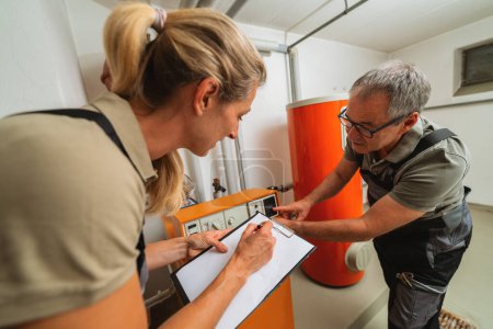 Heizungsingenieure überprüfen eine alte Gasheizung und halten ein Klemmbrett mit Checkliste an einem Heizungsraum in einem Haus. Konzept zum Austausch von Gasheizungen