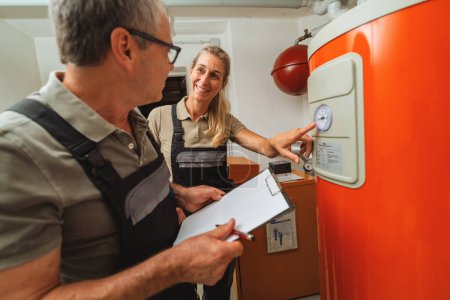 Ein Team von Heizungsingenieuren überprüft ein altes Thermostat für Gasheizungen und hält ein Klemmbrett mit Checkliste an einem Heizungsraum in einem Haus. Konzept zum Austausch von Gasheizungen