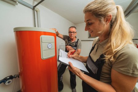 Ein Team von Heizungsingenieuren überprüft eine alte Gasheizung und hält ein Klemmbrett mit Checkliste an einem Heizungsraum in einem Haus. Konzept zum Austausch von Gasheizungen