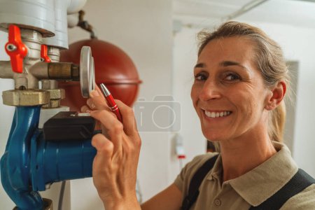 Happy female heating engineer checks gas thermostat at a boiler room with a old gas heating system. Gas heater replacement obligation concept image