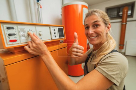 Foto de Happy mujer ingeniero de calefacción muestra el pulgar hacia arriba en una sala de calderas con un viejo sistema de calefacción de gas con lista de verificación en un portapapeles. Imagen del concepto de obligación de sustitución del calentador de gas - Imagen libre de derechos