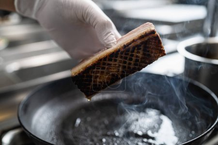 Hand with gloves holding a Crispy pork belly which was roasted in a hot oiled pan at a gas stove in a professional kitchen at a restaurant. Luxury hotel cooking concept image.