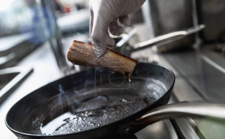 Foto de Mano sosteniendo un vientre de cerdo crujiente que fue asado en una sartén engrasada caliente en una estufa de gas en una cocina profesional en un restaurante. Imagen concepto de cocina de hotel de lujo. - Imagen libre de derechos