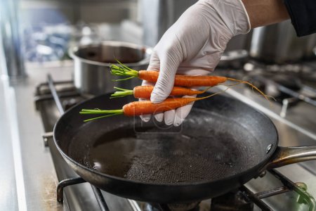 Hand puts fresh carrots into a hot oiled pan at a gas stove in a professional kitchen at a restaurant. Luxury hotel cooking concept image.