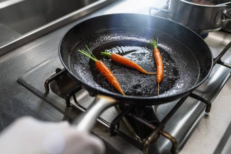 Foto de Mano tirando zanahorias en una sartén engrasada a una estufa de gas en una cocina profesional en un restaurante. Imagen concepto de cocina de hotel de lujo. - Imagen libre de derechos