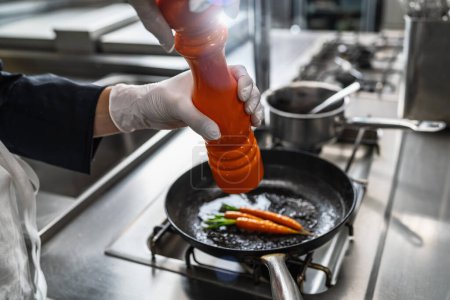 Koch in Hotel- oder Restaurantküche kocht und würzt gebratene Karotten in der Pfanne mit einer Pfeffer- oder Salzmühle. Image der Luxusküche