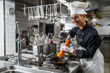 Smiling female chef in hotel or restaurant kitchen cooking and seasoning the food with a pepper or salt mill