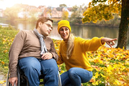 Social service woman shows her friend in a wheelchair something through the Riverside at Park in autumn. Supportive Moments concept image