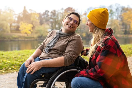 Hombre en silla de ruedas hablando con su cuidadora mujer en un camino mientras camina por el río en otoño. Conexiones alegres concepto de imagen