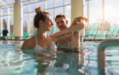 Entrenador masculino que ayuda con el ejercicio de hombro en la piscina del balneario para aeróbicos acuáticos en rehabilitación