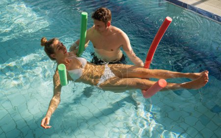 Male trainer using pool noodles to assist a client with rehabilitation exercises in a pool