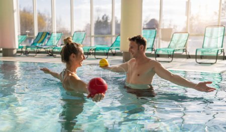 Entrenador masculino realizando terapia acuática con una clienta usando bolas de ejercicio en una piscina soleada