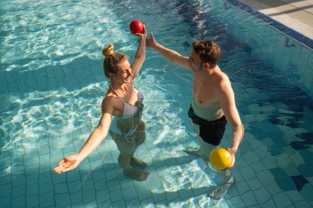 Woman and man in pool lifting colorful exercise balls above water in a sunny rehabilitation session