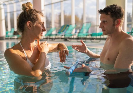 Male trainer explaining exercise to female client in pool, both holding resistance bars, with sunlight