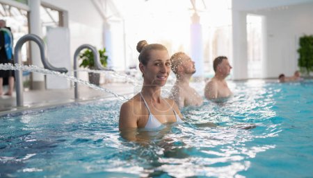 Kurgäste in einem Pool, der von Wasserbächen massiert wird, während eine lächelnde Frau eine Nackenmassage genießt. Image Hotel und Wellness.