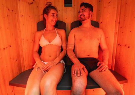 Foto de Pareja sentada en una sauna de infrarrojos, mujer en bikini blanco, hombre en pantalones cortos de baño en un hotel spa - Imagen libre de derechos