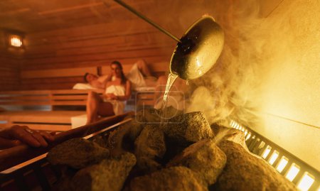 Wasser auf heiße Steine gießen in einer Sauna (lyly), Dampf steigt auf, im Hintergrund entspannen sich zwei Menschen. Wellness Spa Hotel Konzept.
