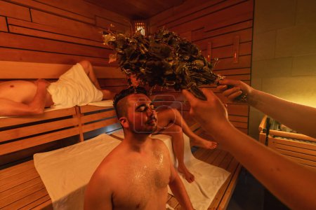 Hombre experimentando un ritual de sauna vihta con otra persona sosteniendo el batidor de abedul, luz cálida en una sauna finlandesa. Wellness Spa Hotel Imagen conservada.