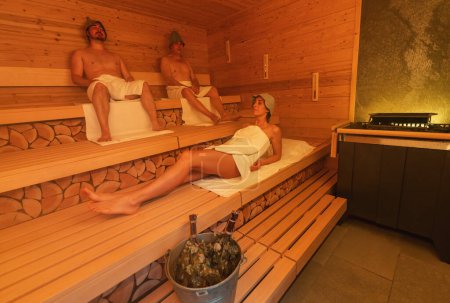 Relaxed individuals in a finnish sauna, two men sitting with felt hats and a woman lying down with a bucket and vihta on the bench