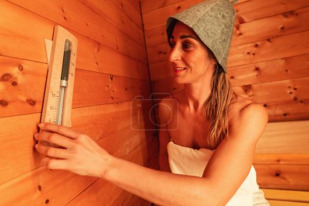 Woman in a sauna wearing a felt hat, smiling while adjusting a wall-mounted thermometer