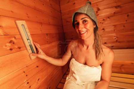 Mujer sonriente en una sauna de acabado que usa un sombrero de fieltro y ajusta un reloj de arena montado en la pared en el hotel spa