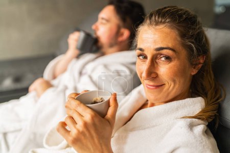Woman with a cup of tea smiling at camera, man in spa robe drinking tea in background at wellness resort