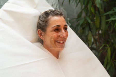 Smiling woman wrapped in a seat steam sauna, with greenery in the background at a wellness hotel