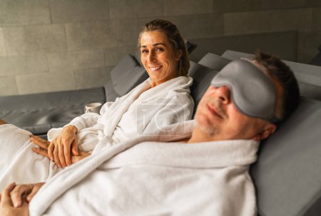 Foto de Mujer en vestido de spa sonriendo, hombre a su lado con máscara de dormir, ambos reclinados - Imagen libre de derechos