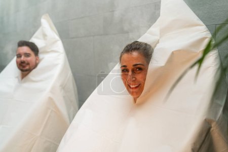 Mujer sonriente y hombre envueltos en bolsas de sauna de vapor caliente térmica en un hotel spa, vapor y plantas alrededor