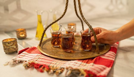 A hand serving Turkish tea on a brass tray with a decorative glass holder and oil bottle in background