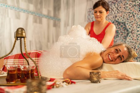 Masseurin trägt Seifenschaum für Hamam auf den Rücken einer entspannten Frau in einem türkischen Bad mit kompliziertem Fliesenhintergrund auf