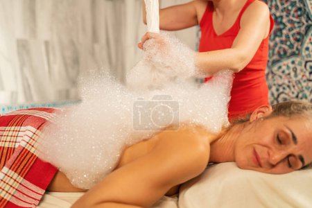 Masseuse applies soap foam for hammam on a relaxed woman's back 