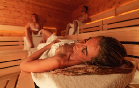 Foto de Personas relajándose en una sauna finlandesa de madera, luz cálida, se centran en la mujer acostada - Imagen libre de derechos