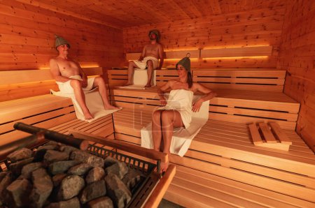 Three people in a sauna wearing finnish felt hats, two men seated and a woman reclining