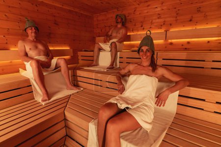 Three people in a sauna wearing finnish felt hats, two men seated and a woman reclining