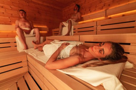 Foto de Mujer reclinada y hombres sentados en una sauna finlandesa de madera, luz ambiental cálida - Imagen libre de derechos