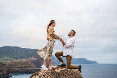 Foto de A young couple stands on a rocky cliff by the ocean, with the man down on one knee proposing to the woman. The ocean stretches out behind them and the sound of waves crashing can be heard in the - Imagen libre de derechos