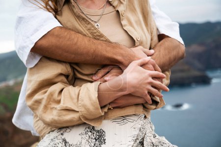 Téléchargez les photos : A close-up of a young couples hands and upper bodies as the man hugs his partner from behind. The womans hands are resting on top of his, and their fingers are entwined. The setting appears to be a - en image libre de droit