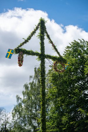 Image of A pole and flag against green trees and blue sky. A maypole decorated, covered in flowers and leaves.