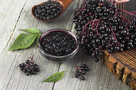 Delicious homemade black elderberry syrup in glass jar and bunches of black elderberry with green leaves on wooden desk.