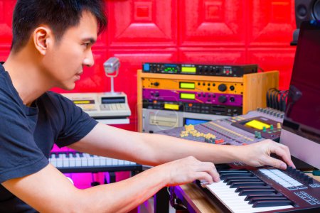 asiatischer gutaussehender professioneller Musikproduzent, der im Heimstudio einen Hit per Midi-Keyboard auf dem Computer arrangiert